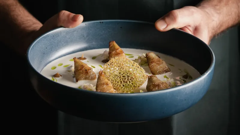 Chef holding a blue ceramic bowl with creamy soup, roasted leeks, olive oil drops, and a lacy cheese crisp garnish
