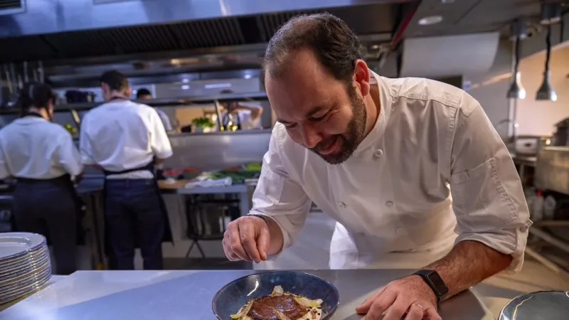 Smiling chef in white jacket carefully finishing a dish in a blue bowl on a steel counter in a busy restaurant kitchen