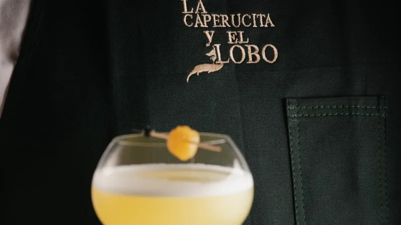 Dark apron embroidered with La Caperucita y El Lobo logo, with a frothy yellow cocktail garnished with a cherry in front