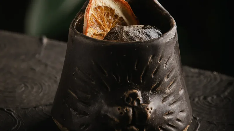 Dark textured ceramic cup with wolf face relief holding a cocktail with ice and a dried orange slice garnish