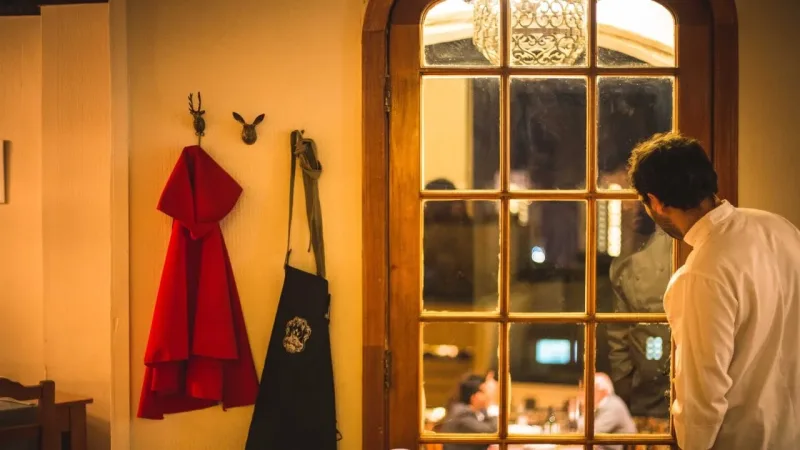 Chef in white jacket looking through a glass-paned door at dining guests, with red and black aprons hanging on wall hooks