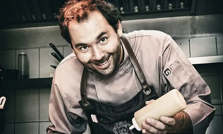 Smiling male chef in apron holding a squeeze bottle over a plated dish in a restaurant kitchen