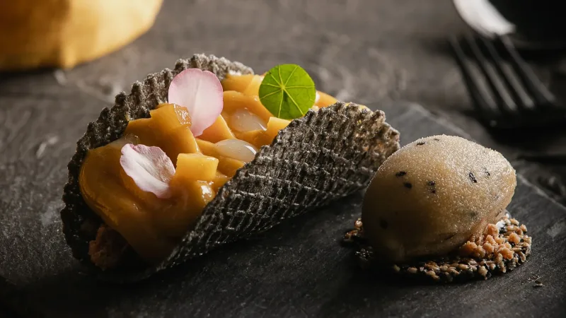 Elegant dark waffle tart filled with caramel fruit compote, edible flower petals, and a scoop of grey ice cream on a slate