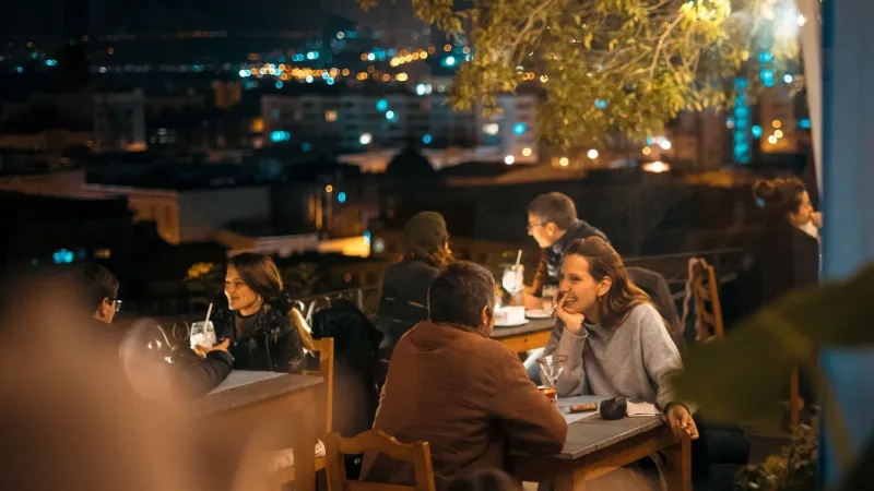 People dining and socializing at an outdoor rooftop terrace bar at night with a glowing city skyline in the background