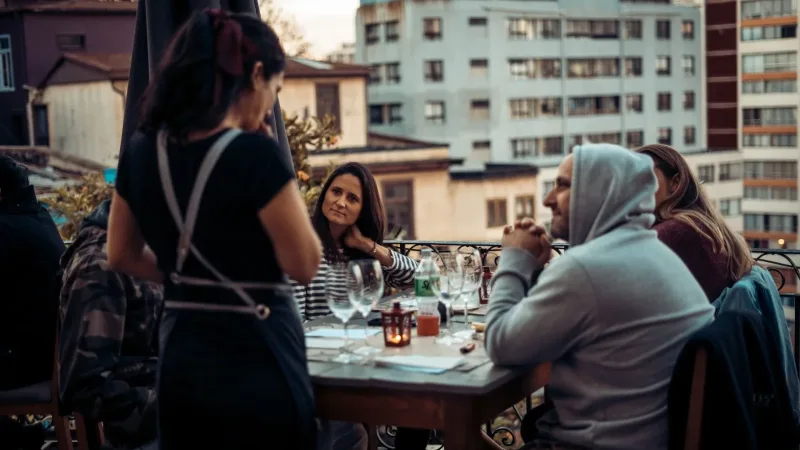 A waitress takes an order from a group of people dining at an outdoor rooftop restaurant with city buildings in the