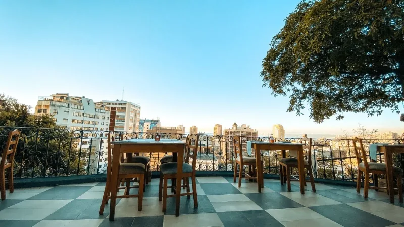 Outdoor rooftop terrace with wooden tables and chairs, checkered tile floor, wrought iron railing, and city skyline view