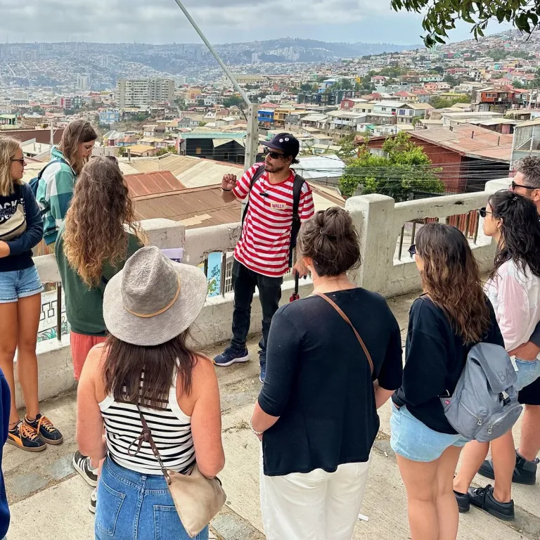 Guía turístico con polera a rayas rojas lidera un grupo de turistas en un mirador con vista panorámica de Valparaíso.